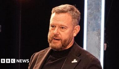 Mark Ashton has short brown hair and is wearing a brown suit jacket over a black rollneck top. He is speaking on stage.