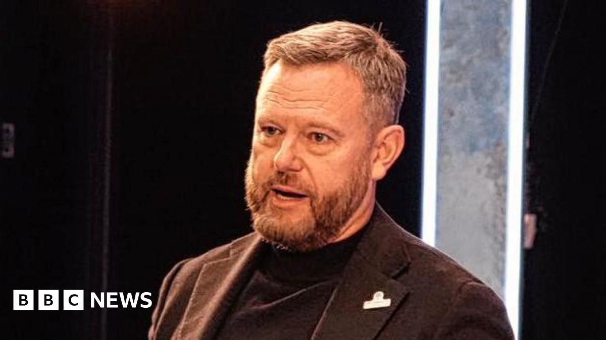 Mark Ashton has short brown hair and is wearing a brown suit jacket over a black rollneck top. He is speaking on stage.