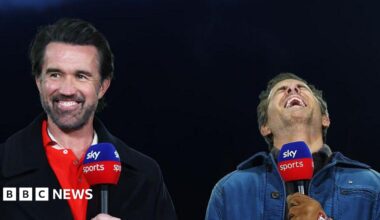 Rob Mac and Ryan Reynolds holding microphones and laughing as they stand against a dark backdrop. Rob wears a red shirt with a black coat, while Ryan wears a blue jacket and brown gloves