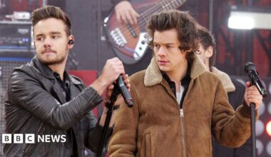 Liam Payne and Harry Styles performing together on stage. The pair both wear black and white suits and there is a band behind them