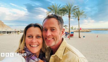 A couple on a beach. It is sunny and the sand is white. Behind them are some trees.