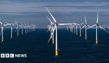 Two wind turbines at sea, one close to the camera showing only the turbine and part of the blades. The other is seen in the distance, with a yellow marking at the base of the white tower that supports the turbine. It is a clear day with calm waters.