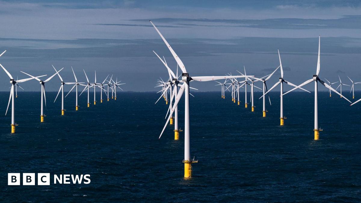 Two wind turbines at sea, one close to the camera showing only the turbine and part of the blades. The other is seen in the distance, with a yellow marking at the base of the white tower that supports the turbine. It is a clear day with calm waters.
