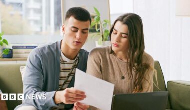 A young couple sit on a sofa in their living room looking at a bill with concern