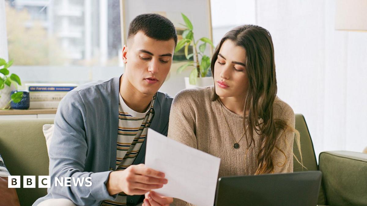 A young couple sit on a sofa in their living room looking at a bill with concern