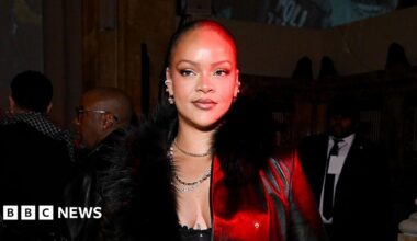 Rihanna has short hair wearing a black furry coast with a red light cast on her face