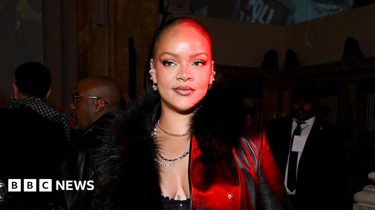 Rihanna has short hair wearing a black furry coast with a red light cast on her face