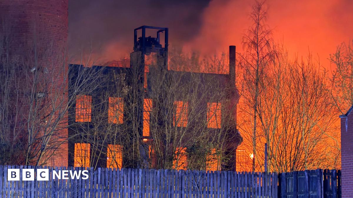 A large blaze at night in a mill building. The sky is lit up orange. Wooden fencing is in front of the building.