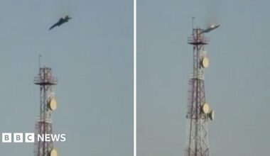 A man wearing a red hoodie in Tehran  is seen from behind watching an explosion and smoke rising in the distance through binoculars