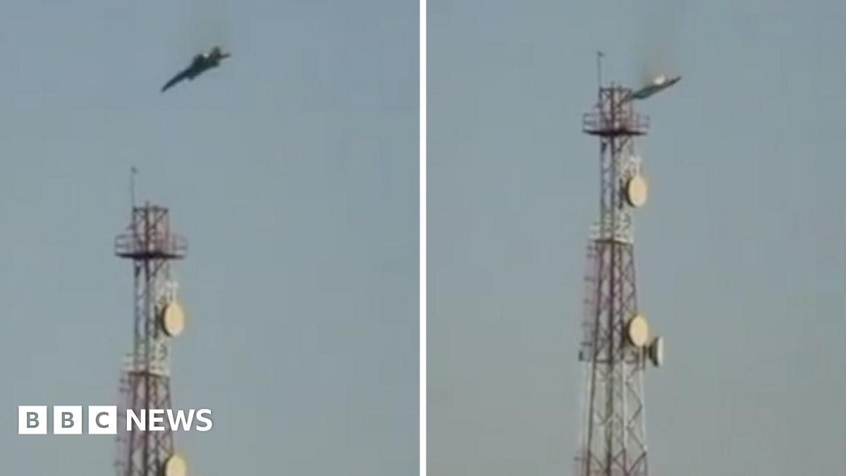 A man wearing a red hoodie in Tehran  is seen from behind watching an explosion and smoke rising in the distance through binoculars