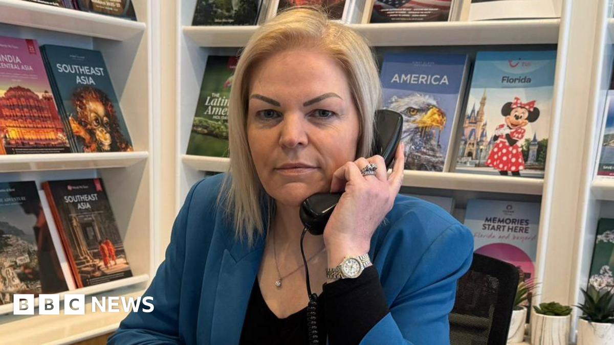 Linda Hill Miller, a woman with blonde, shoulder length hair. She is wearing a black top with a blue blazer and holds a phone to her ear. In the background there are travel brochures for different countries on shelves.
