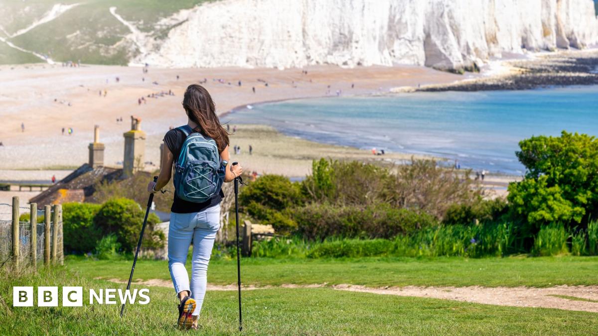 A wide, aerial shot of a part of King Charles III England Coast Path in Hampshire shows four people walking on a path by sea shoreline on a sunny day. The stretch of land between the sea and the path looks muddy and is covered in green moss and low grass. On the other side of the path, there is brown grassland with a few trees with a green field in the background. In the middle of the photo, there is a stone building and a round tower.