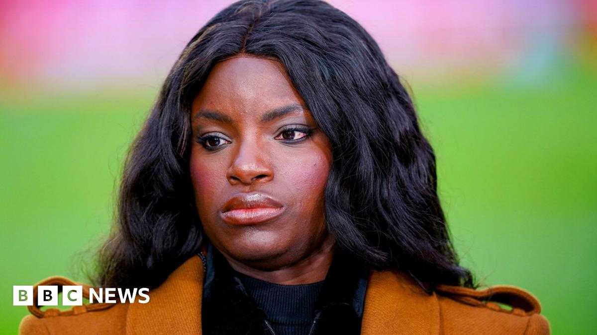 Eniola Aluko looking off-camera. She has long black hair and is wearing an orange jacket.