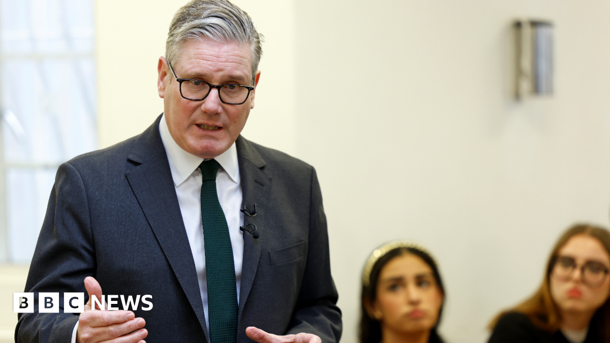Keir Starmer, in a suit and tie,  speaking while two women in the background watch on