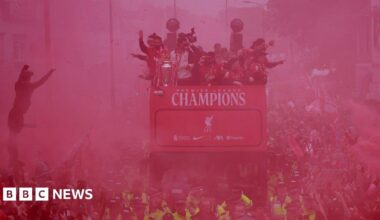 Liverpool FC parade bus passing through a large crowd - the gathered people are obscured by a haze of red smoke.