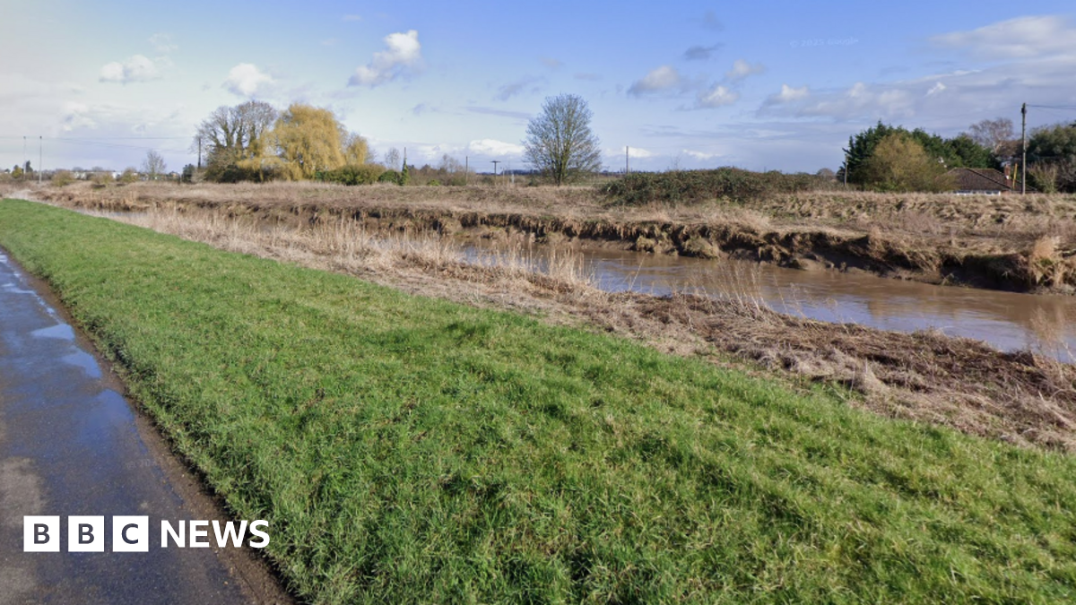 A river running next to a rural single-carriageway road. A short grass verge separates the road from the water.