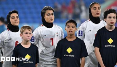 Three women wear white football jerseys and black headscarves while three boys in black t-shirts stand in front of them