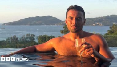 Jordan Wright holding a wine glass at the edge of an infinity pool. He has brown hair and stubble and the sun is shining on him. Tall green hills and the sea are pictured behind him.