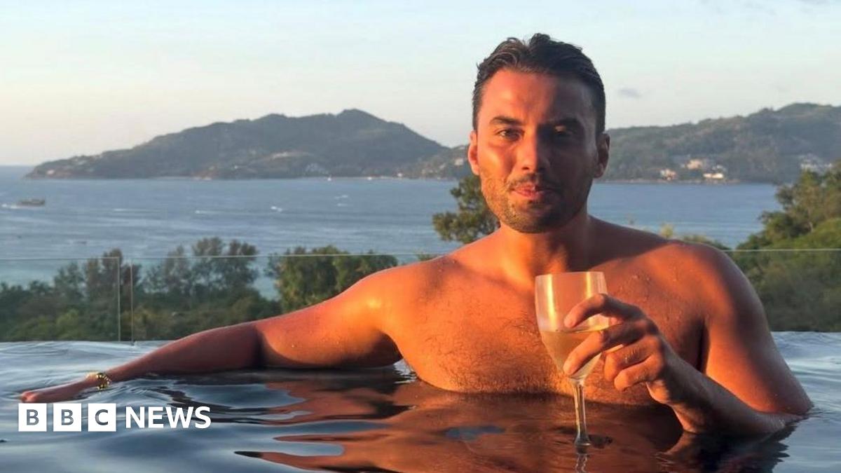 Jordan Wright holding a wine glass at the edge of an infinity pool. He has brown hair and stubble and the sun is shining on him. Tall green hills and the sea are pictured behind him.