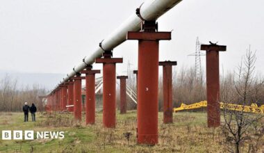 Men stand near the Druzhba crude oil pipeline near Styri, Ukraine in 2009. The pipeline is situated on large orange pillars in a field.