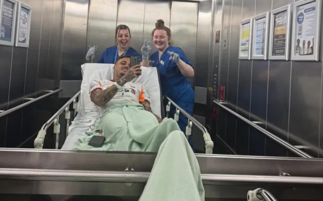 Noa Lang takes a selfie from a hospital bed, surrounded by two smiling nurses in an elevator.
