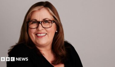 Mid-shot of Liza Tarbuck wearing a black top, leopard print frames and wearing streaks of blonde in her long flowing hair.