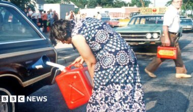 In a photo from the 1970s, a woman in a blue and white dress uses a red jerry can to pour petrol into her brown car. In the background, a man walks behind her car with another jerry can, while a large group of people queue.