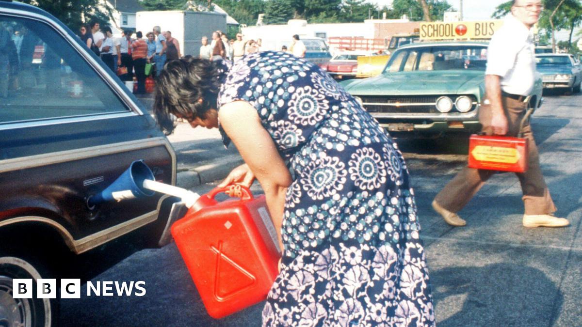 In a photo from the 1970s, a woman in a blue and white dress uses a red jerry can to pour petrol into her brown car. In the background, a man walks behind her car with another jerry can, while a large group of people queue.