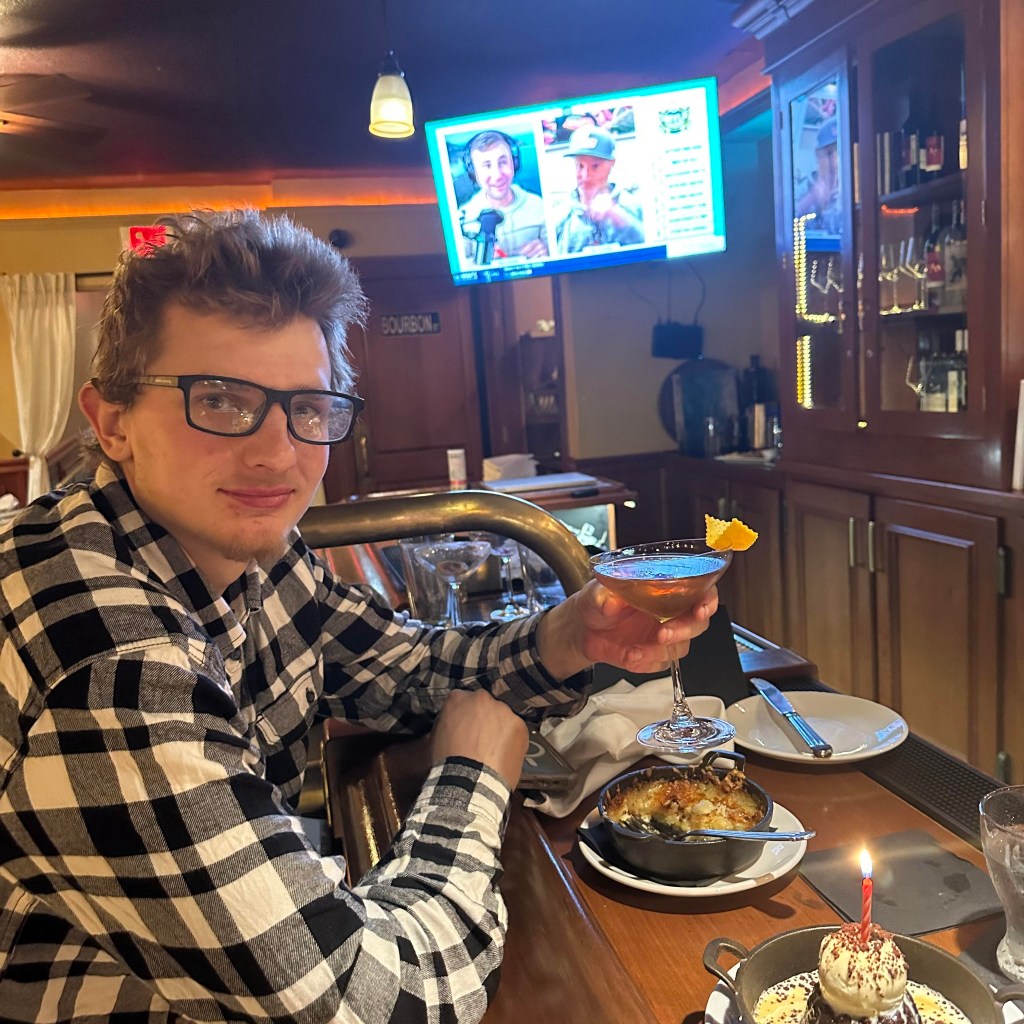 Ben Fowler holding a cocktail at a bar with food and a lit candle in front of him.