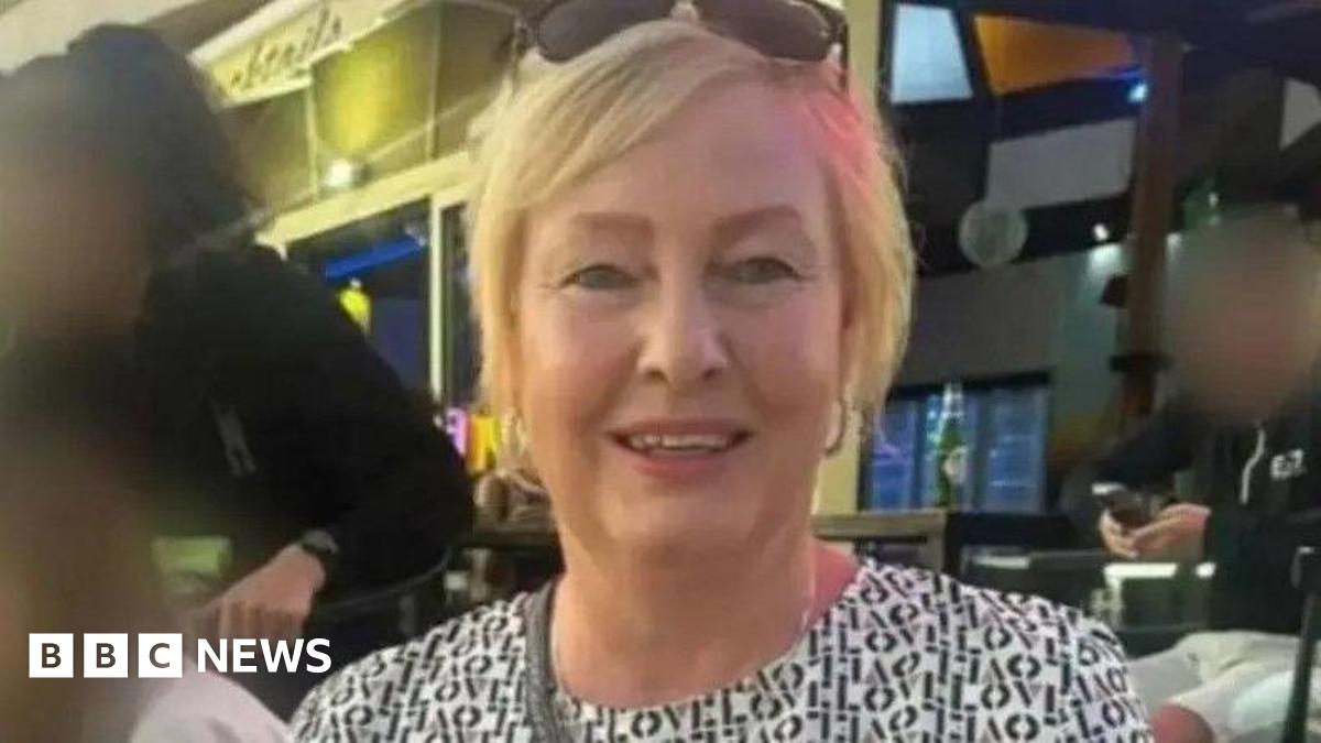 A woman in her fifties with blonde hair. She is wearing sunglasses on her head and appears to be sat in a bar. People behind her have their faces blurred.