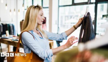 A woman with long blond hair looks at a top in a clothes shop. The woman is wearing a light blue top and has a light brown bag on her shoulder