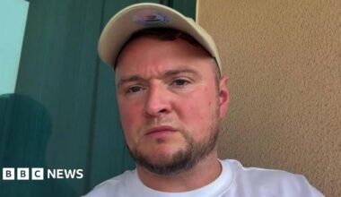 A man with short brown hair sitting in a hotel room looks into a camera