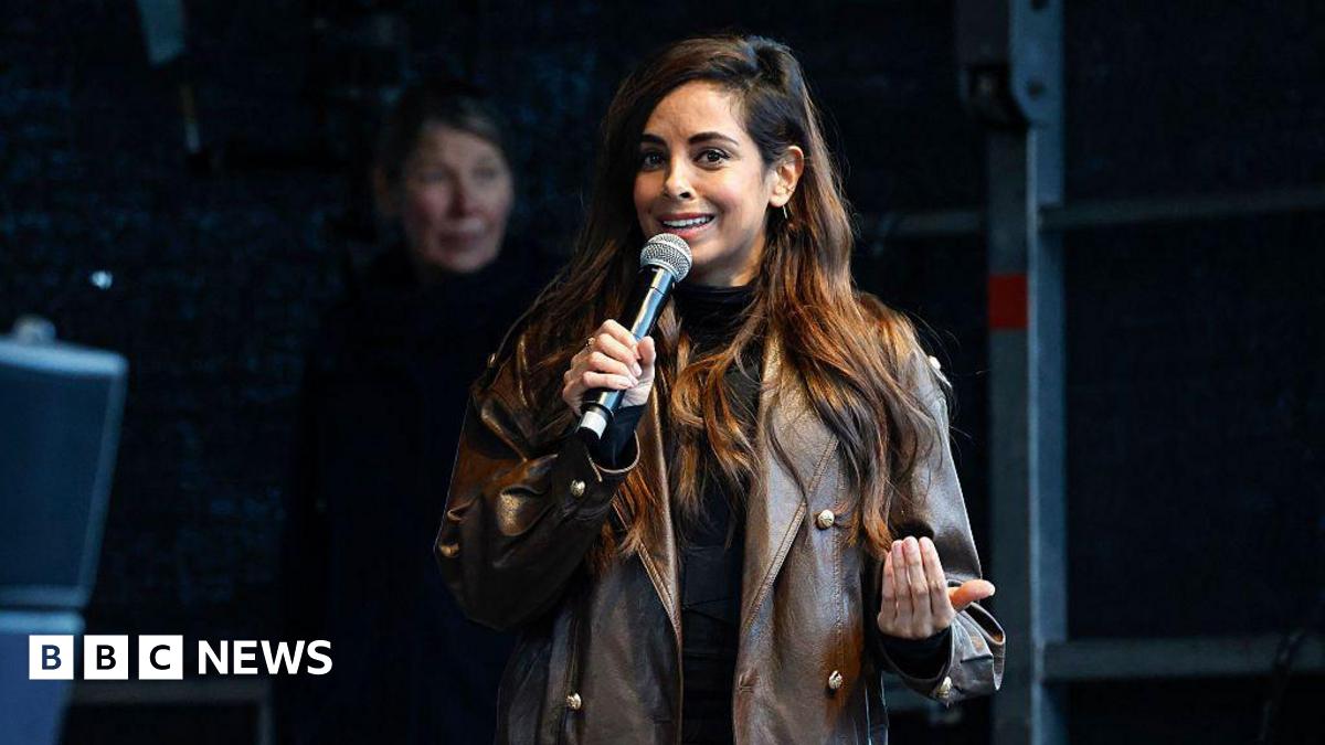 A woman in a brown leather jacket and brown hair holds a microphone