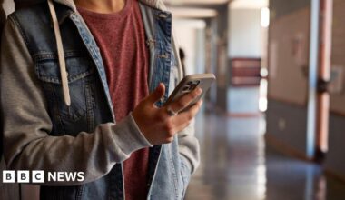 A close-up of an anonymous young person looking at a phone in a school corridor. They have a denim blue and grey jacket and a red T-shirt underneath, with a ring on the finger.