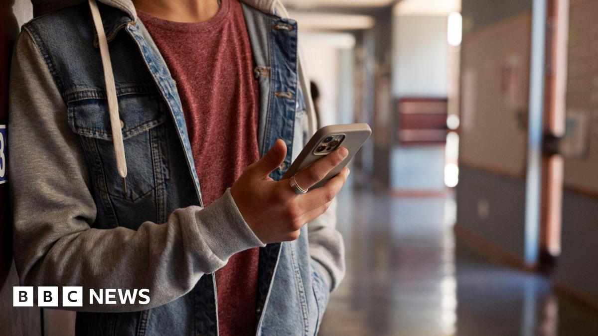 A close-up of an anonymous young person looking at a phone in a school corridor. They have a denim blue and grey jacket and a red T-shirt underneath, with a ring on the finger.