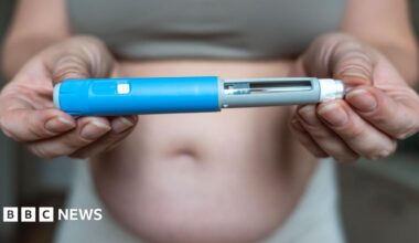 A person holding an injector pen for a weight-loss drug - the pen is in focus at the foreground of the image, held at either end by both hands of the person. To the left, the pen has a blue outer shell, with it then tapering to the right into a grey section, with a see-through strip, to the sharp point of the needle. The person in the background appears to be a woman, we only see their middle third. They are wearing a top that cuts off at the belly, showing the person is overweight.