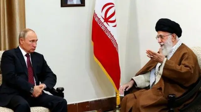 Russian President Vladimir Putin, wearing a black suit and red tie, is having a sit down conversation with Iranian Supreme Leader Ayatollah Ali Khamenei, to his right. The Iranian flag can be seen at the back, between the two men.