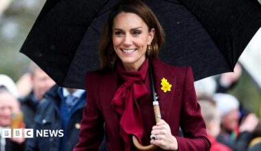 Catherine wearing burgundy with a yellow daffodil pinned to her blazer, holding a black umbrella, as people take pictures in the background