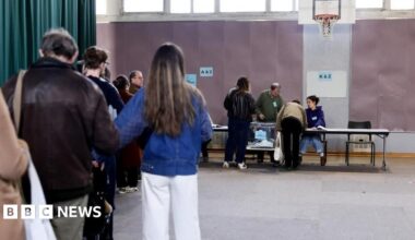 Voters queue inside a polling station at the far end of which two officials at a long table face two of the voters about to retrieve their ballots