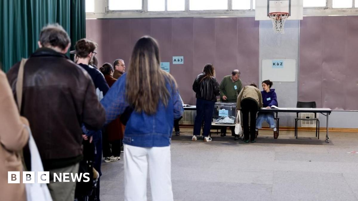 Voters queue inside a polling station at the far end of which two officials at a long table face two of the voters about to retrieve their ballots