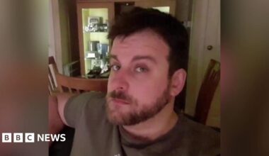 A man with brown hair, a beard and moustache looking at the camera. He is wearing a brown t-shirt. Behind him there are three wooden chairs and a cabinet.