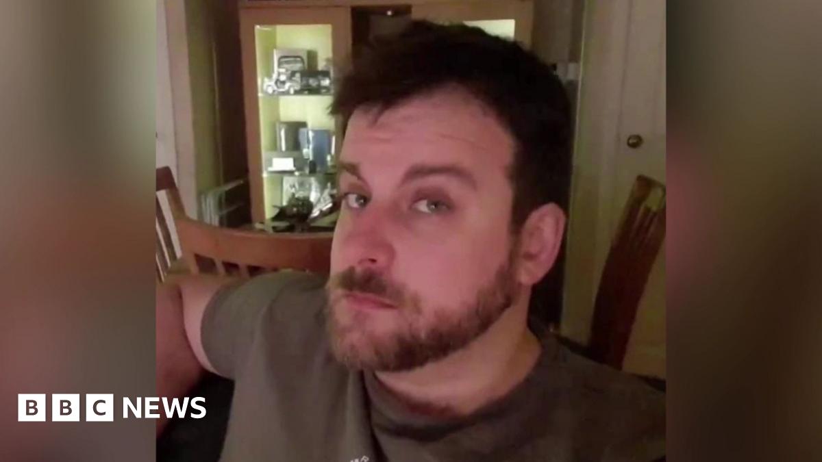 A man with brown hair, a beard and moustache looking at the camera. He is wearing a brown t-shirt. Behind him there are three wooden chairs and a cabinet.