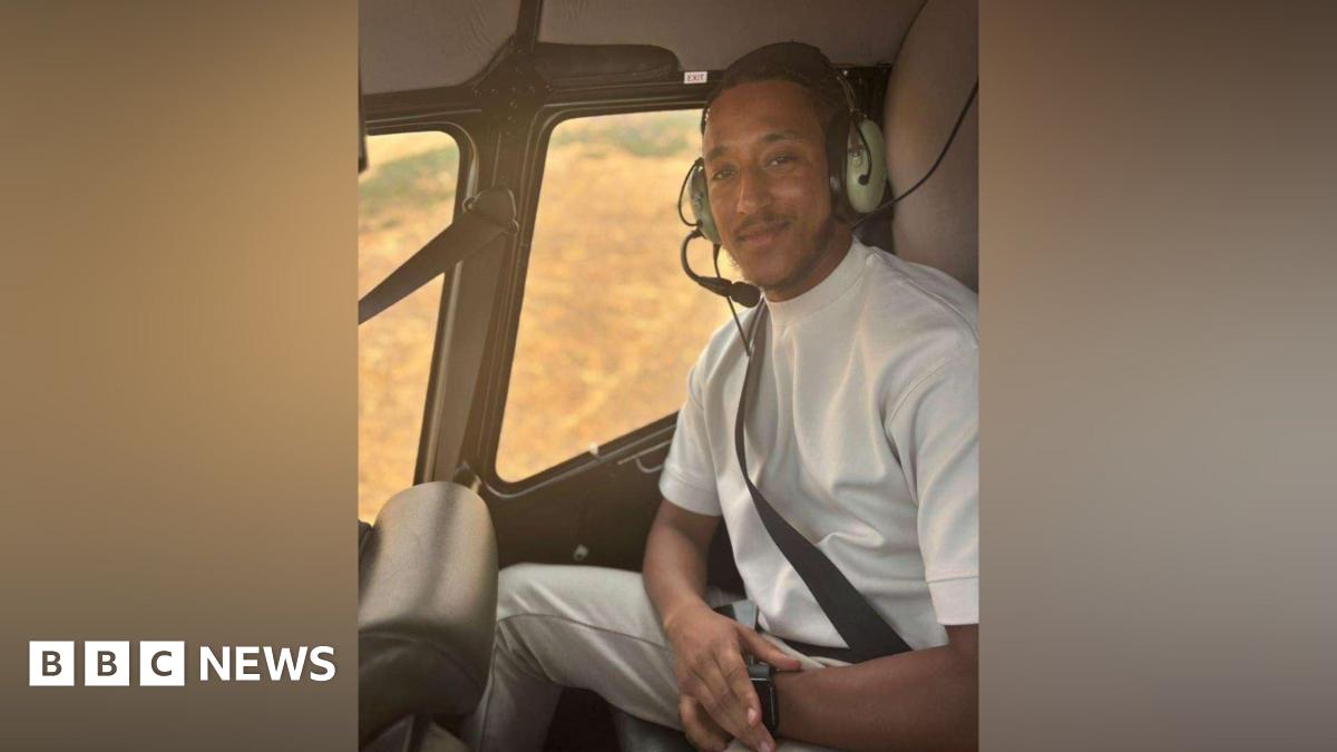 A man in a white shirt sits in a helicopter with a headset on. He smiles at the camera.