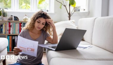 A woman has her hand pressed against her head as she sits on the floor with her laptop on the sofa looking at her energy bill on a piece of A4 paper
