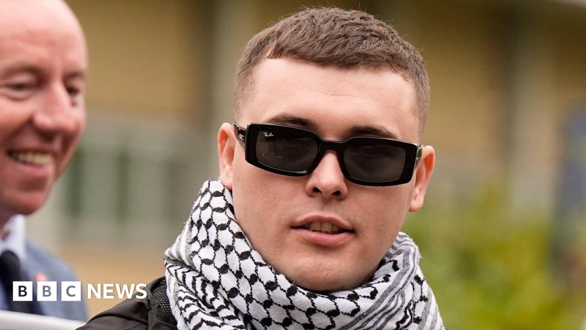 A close-up image of Liam Óg Ó hAnnaidh outside court. He is a young man wearing a pair of black RayBan sunglasses. He has a white and black keffiyah around his neck. He is looking off to the side.