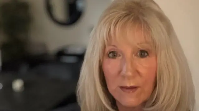 A woman with blonde hair and a pearl necklace is looking at the camera, sitting in front of a blurred background which appears to be a dining room.