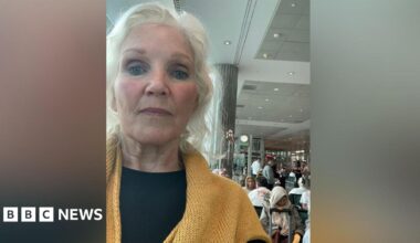 Lenise Blom, who is wearing a yellow jumper and black shirt takes a selfie while stood inside the terminal at Dubai Airport. Dozens of people are sat behind her. She is a woman in her 70s who has wavy white hair.