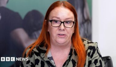 Charlotte Hennessy, daughter of Hillsborough victim Jimmy Hennessy, has long red hair and is photographed wearing black glasses and an olive green and black patterned top. She is speaking during a press conference.