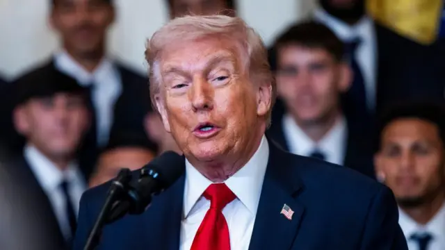 US President Donald Trump speaking into a microphone while wearing a navy blue suit with a red tie and an American flag pinned to his suit.