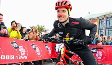 Radio 1 DJ Greg James is on his red bike on the starting line runway. Either side is a metal barrier lined with people waving. He is wearing all black cycling outfit and a red helmet and a red nose.